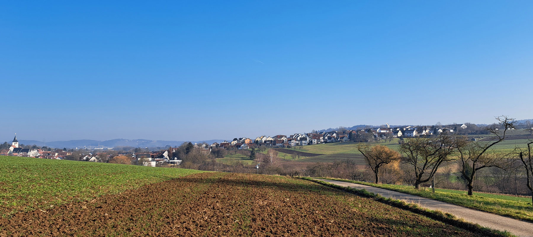 Weitblick über Felder und Wiesen auf die Ortschaft mit Kirche und Häusern unter klarem blauem Himmel.