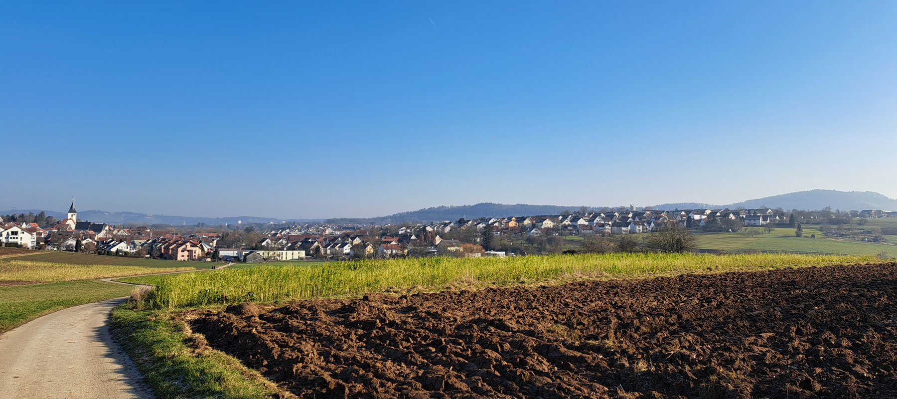 Landschaftsaufnahme mit Feldweg, Ackerland und Blick auf die Ortschaft mit Kirche unter klarem blauen Himmel.